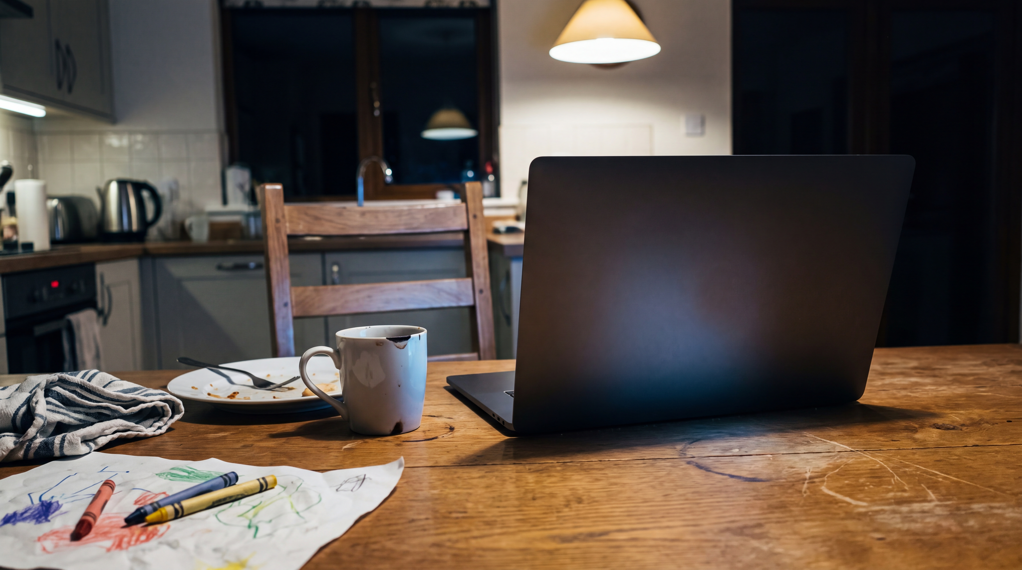 An open laptop on a kitchen table at night with its screen turned away from the camera, beside a scraped dinner plate and fork, a stained mug, a folded dish towel, and two crayons lying on a child's scribbled drawing, an empty chair pushed back behind it.